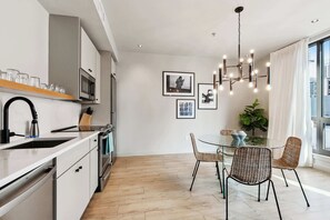 Eine moderne Küche mit weißen Schränken, einer Edelstahl-Mikrowelle und einem Holzregal. Ein Essbereich mit Korbstühlen und einem Glastisch.