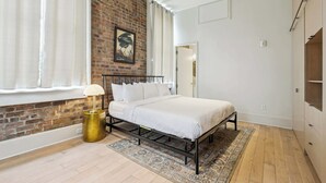 Une chambre avec un mur en briques, un lit, une fenêtre avec des rideaux, une lampe et un tapis.