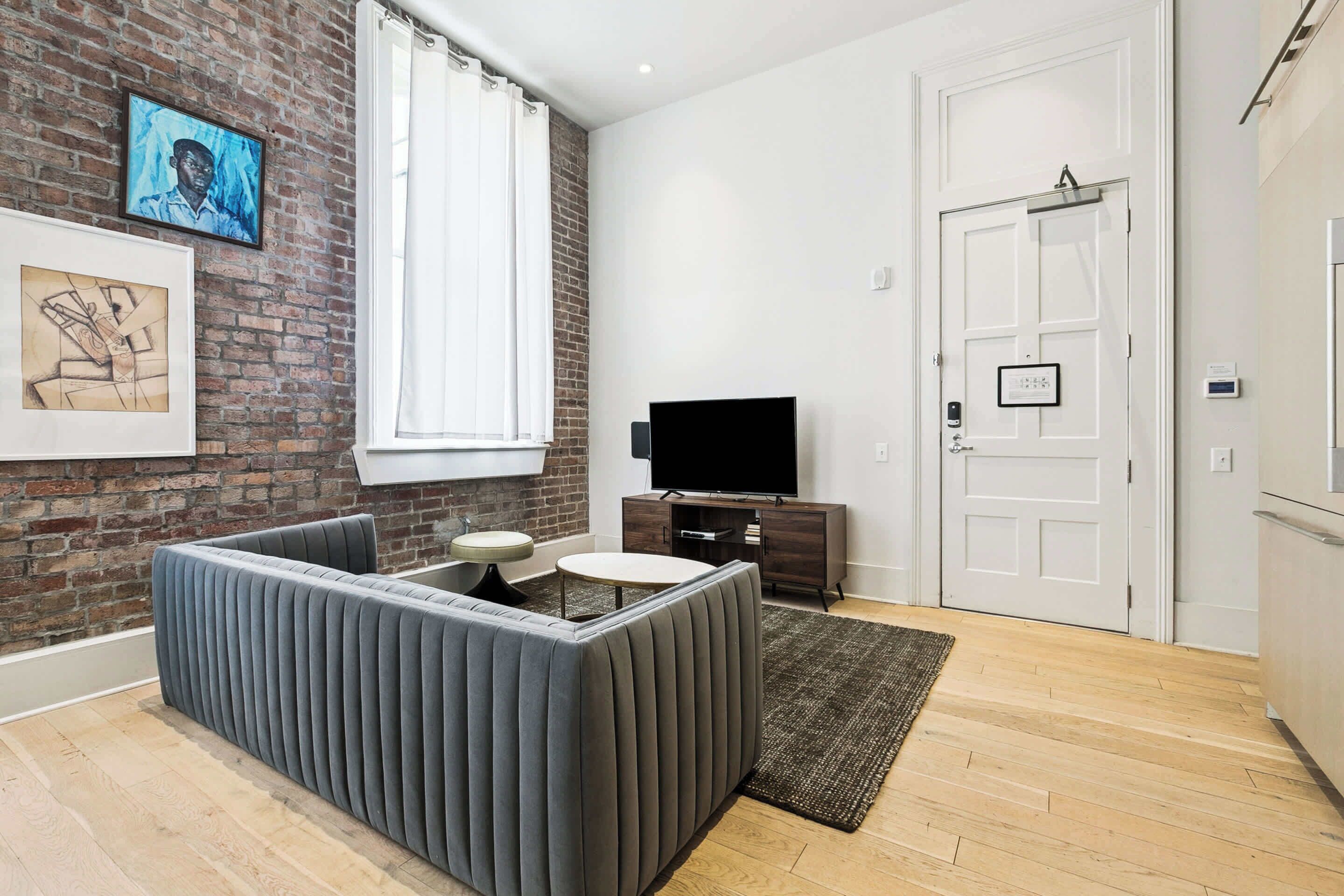 A modern living room with a brick wall, a flat-screen TV, a grey sofa, a small round table, and a white door.