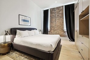 Une chambre à coucher moderne avec un grand lit, une table de chevet, une lampe, un tableau encadré au mur et un mur de briques visible à travers la fenêtre.
