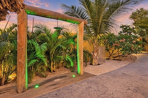 Eine hölzerne Pergola mit grüner Beleuchtung, umgeben von tropischen Pflanzen und einem Pfad.