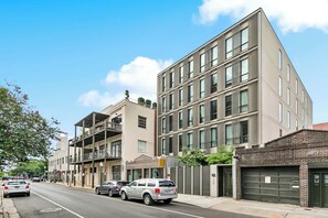 Une rue moderne en milieu urbain, bordée d’immeubles à plusieurs étages, avec des voitures garées et un ciel dégagé.