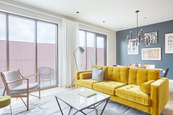 Un salon spacieux avec un canapé jaune, une table basse en verre et une grande fenêtre donnant sur un immeuble.