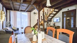 Zona de menjador acollidora amb taula de fusta, cadires i un candelabrum. Hi ha una televisió penjada a la paret i una finestra amb cortines.