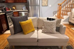 Un salon avec un canapé beige, un coin cuisine doté d’un réfrigérateur en acier inoxydable, de meubles de rangement en bois et d’un escalier.