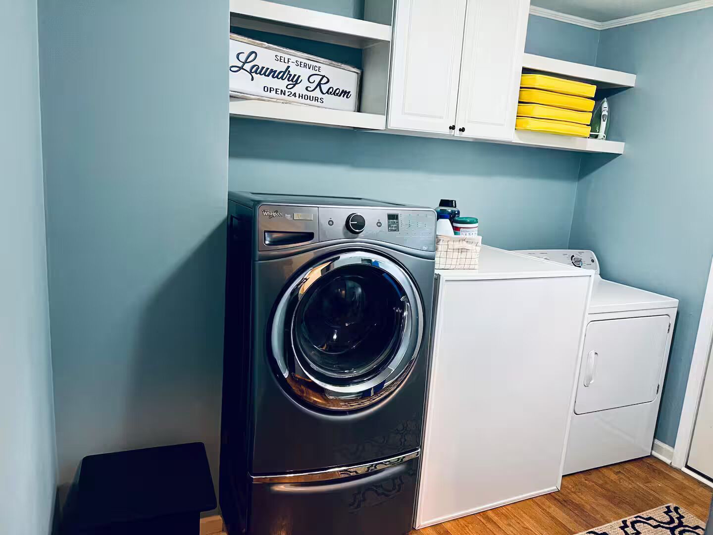 A laundry room with a front-loading washer and dryer, a shelf with laundry supplies, and a sign indicating it's self-service and open 24 hours.