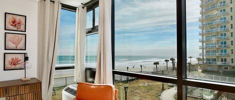Habitación con un ventanal grande que da a una playa y un edificio alto. Se observa un armario de madera, una silla naranja y una pequeña mesa redonda.