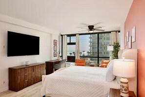 Habitación de hotel con cama, televisión, ventilador de techo, escritorio y vista de la ciudad a través de amplias ventanas.