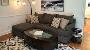 Una sala de estar con un sofá modular, una mesa de centro de vidrio, una alfombra estampada, luces fijadas a la pared y un cuadro de olas.