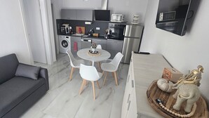 Ett kompakt vardagsrum med en grå soffa, ett runt matbord med vita stolar, ett köksområde med moderna vitvaror och en dekorativ elefantfigur.