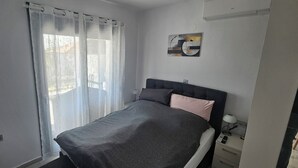 Ett sovrum med en säng, ett sängbord med en lampa, en väggmonterad luftkonditioneringsanläggning och ett fönster med tunna, genomskinliga gardiner.
