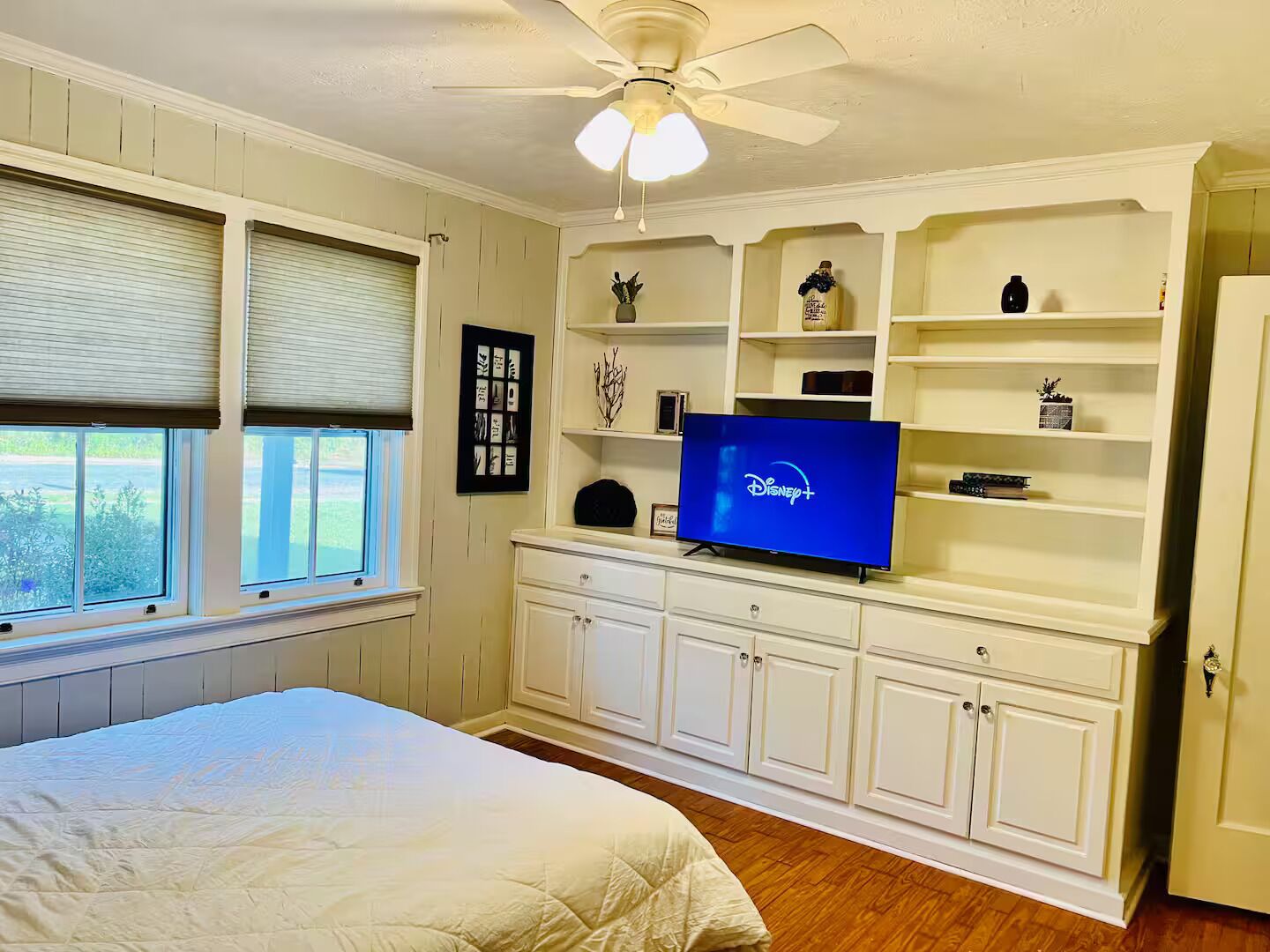 Une chambre à coucher avec un lit, un téléviseur posé sur un meuble blanc, un ventilateur de plafond et une fenêtre avec des stores.