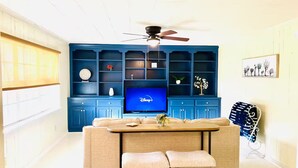 Een woonkamer met een blauwe entertainmentkast, een plafondventilator, een televisie, een bank en een eettafel met twee stoelen.