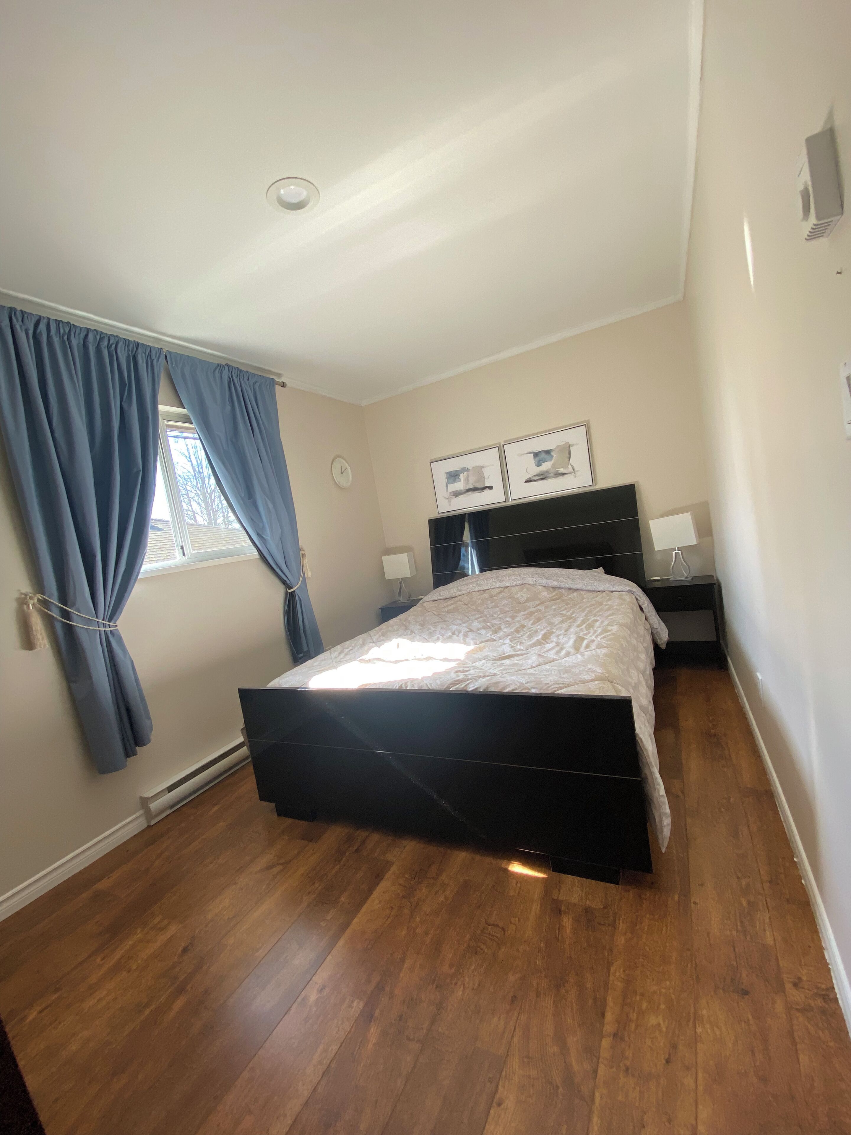 A bedroom with a bed, a nightstand, a dresser, a window with curtains, and a wall clock.