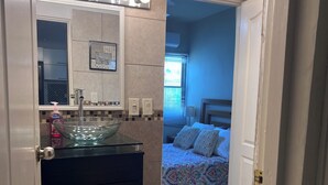Un bany amb un lavabo, un mirall i il·luminació. A l'habitació propera hi ha un llit amb roba de llit estampada.