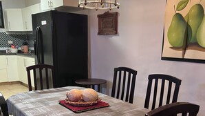 Una zona de menjador amb una taula preparada per a quatre, envoltat de cadires de fusta. Es veu un frigorífic negre al fons, i un quadre de peres penja a la paret.