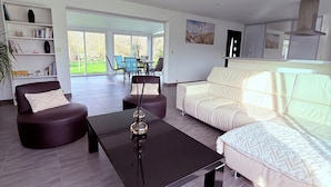 Een moderne woonkamer met een bank, fauteuil en salontafel. Achtergrond toont een boekenkast en een eethoek.