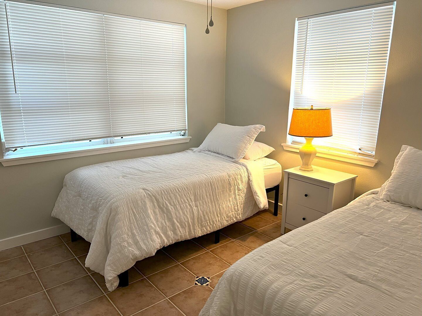 A bedroom with two beds, a bedside table with a lamp, and windows with blinds.