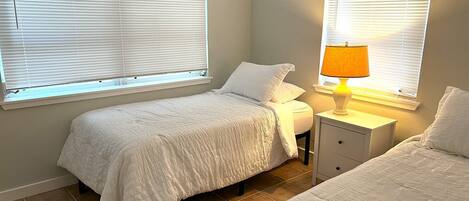 A bedroom with two beds, a bedside table with a lamp, and windows with blinds.
