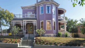 Une maison à deux étages avec une véranda qui fait le tour de la maison, une entrée principale avec une porte, et un petit jardin devant la maison avec un arbre et des arbustes.