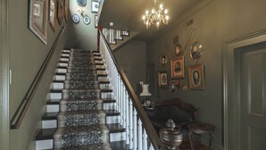 Een trap met een gestreept tapijt en witte spijlen, die leidt naar een kamer met ingelijste foto's en een kroonluchter.