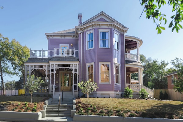 Une maison à deux étages avec une véranda qui fait le tour de la maison, une entrée principale avec une porte, et un petit jardin devant la maison avec un arbre et des arbustes.