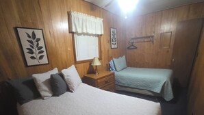 Una camera da letto con un letto grande, un letto più piccolo, pareti in legno, una ventola a soffitto, una finestra con persiane e un attaccapanni.