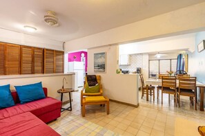 Ein kompakter Wohnbereich mit einem roten Sofa, einem gelben Sessel, einem Essbereich mit Tisch und Stühlen und einer Küchenzeile im Hintergrund.