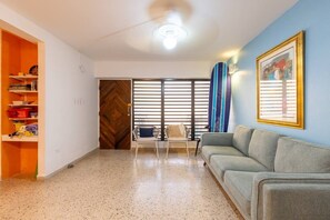 Ein Wohnzimmer mit einer Couch, einem Sessel, einer Holztür, einem Fenster mit Jalousien und einem Surfbrett.