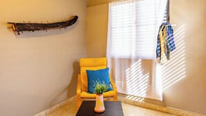 Een hoekzaak met een gele fauteuil, een vaas met een plant, een houten wanddecoratie en een raam met zonwering.