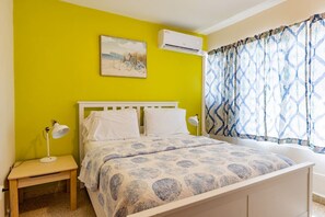 A room with a bed, a bedside table with a lamp, a wall-mounted air conditioner, and a window with patterned curtains. - Vilomar B7 Walking distance to beach in Luquillo (Luquillo)