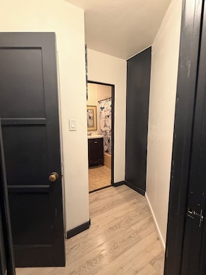 A narrow hallway with a wooden floor, a black door on the left, and a bathroom visible in the background.