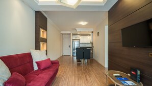 Un salon moderne avec un canapé rouge, un téléviseur à écran plat fixé sur un mur en bois foncé, et une cuisine visible au fond.