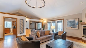Een ruime woonkamer met een grote bank, een salontafel en een open haard. Achtergrond is een keuken met houten kastjes en een vaatwasser.