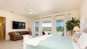 A bedroom with a large bed, a ceiling fan, a TV, a brown leather sofa, and a view of the outdoors. - Temecula Wine Country Hilltop Estate – 360° Views & Heavenly Sunsets (Temecula)