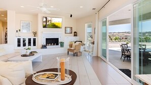 A modern living room with a fireplace, a ceiling fan, and a large window overlooking a scenic landscape. - Temecula Wine Country Hilltop Estate – 360° Views & Heavenly Sunsets (Temecula)