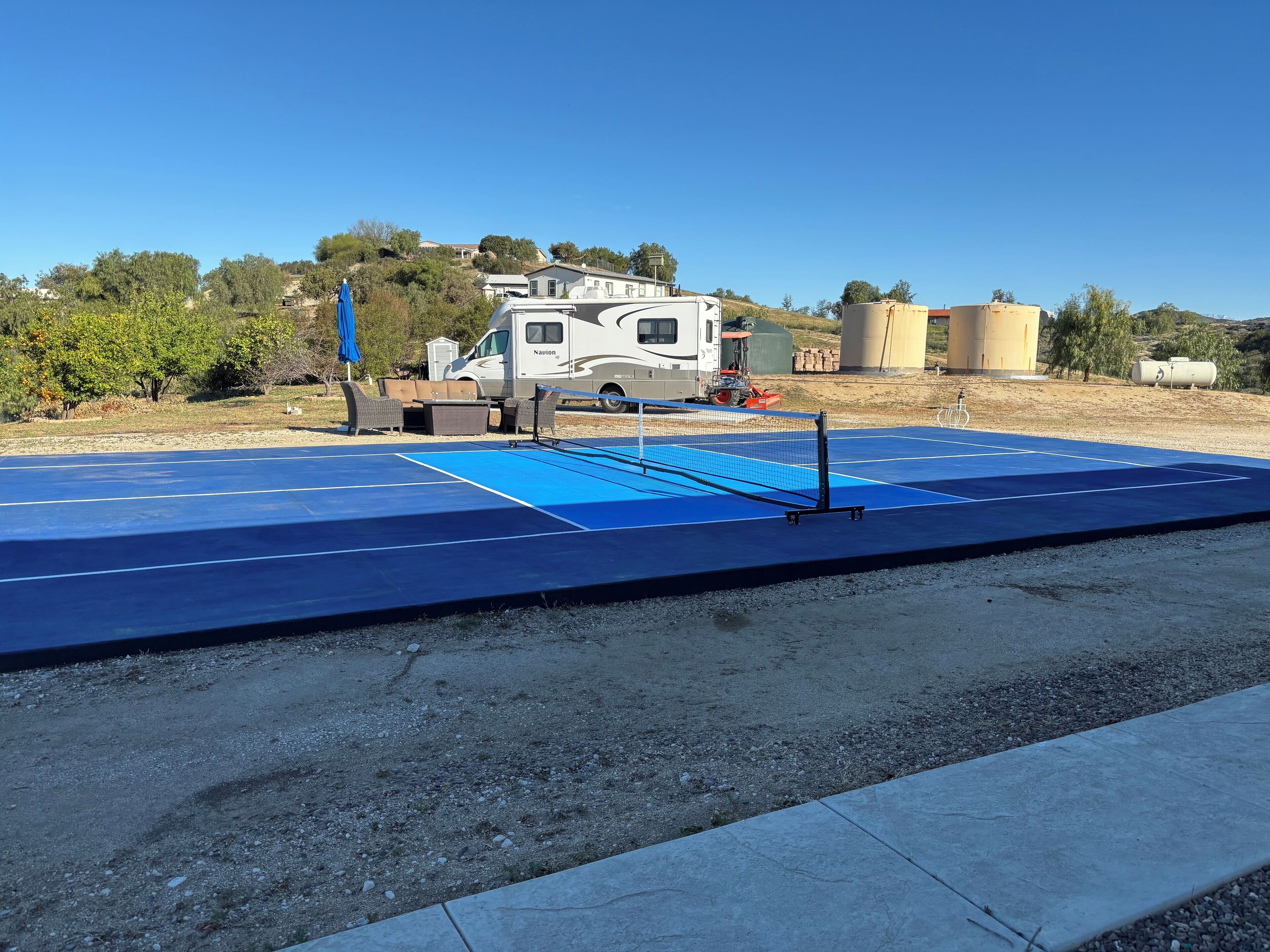 A blue tennis court with a net, surrounded by gravel and a concrete edge.