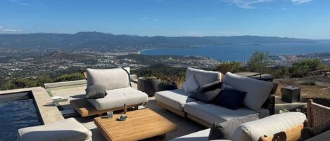 Een terras met buitenmeubilair, een zwembad en een mooi uitzicht op een kuststad en bergen.