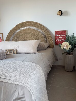 A bedroom with a wicker headboard, a bed with white bedding, a 'Welcome to the House' sign, a wooden sign with 'ZEN,' and a vase with flowers. - L'écrin de Paradis (ALATA)