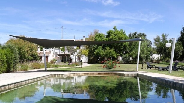 A swimming pool with a shaded area, surrounded by greenery and a building in the background. - Cozy 1-Bedroom Apartment with Terrace and Pool in Bensafrim (Bensafrim)