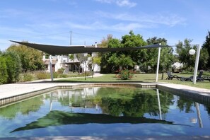 Una piscina con una zona sombreada, rodeada de vegetación y un edificio al fondo.