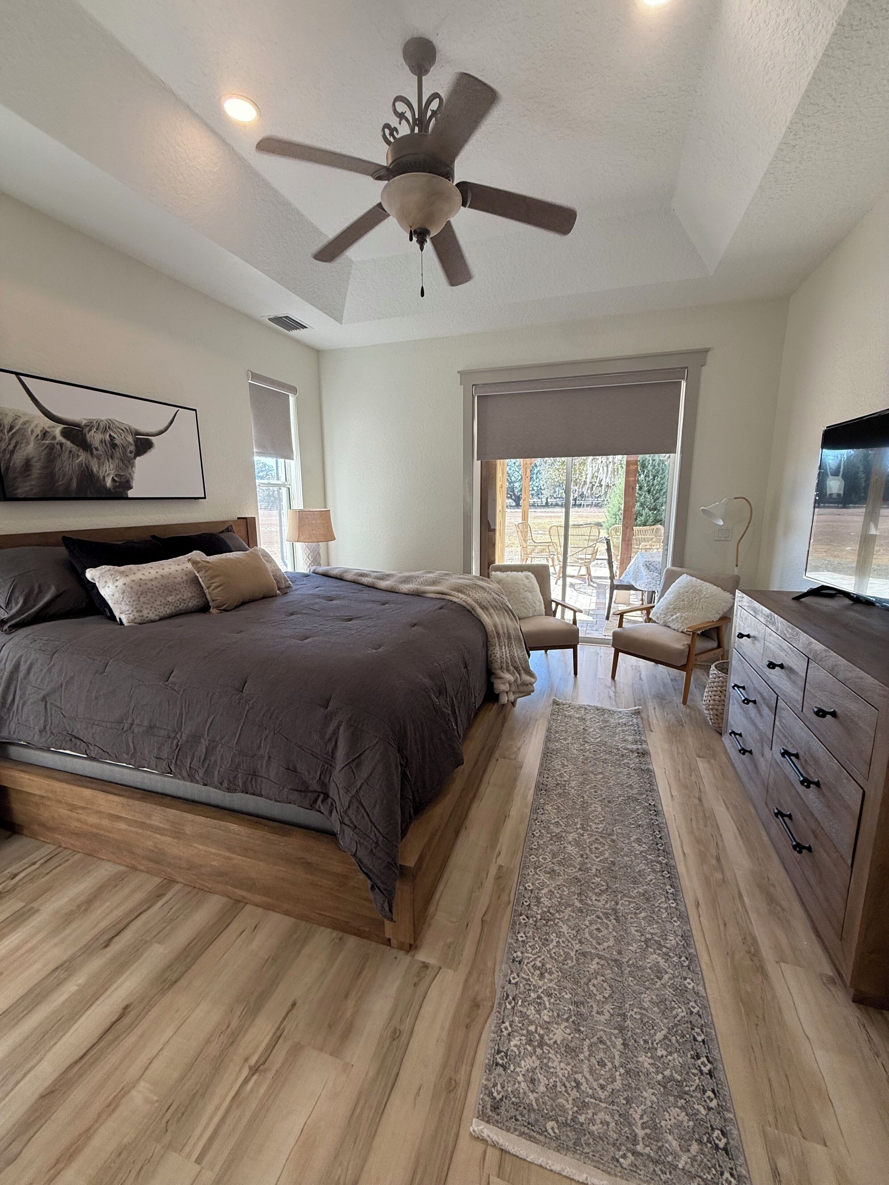 A bedroom with a large bed, a ceiling fan, a wooden dresser, and a view of the outdoors through a sliding door.