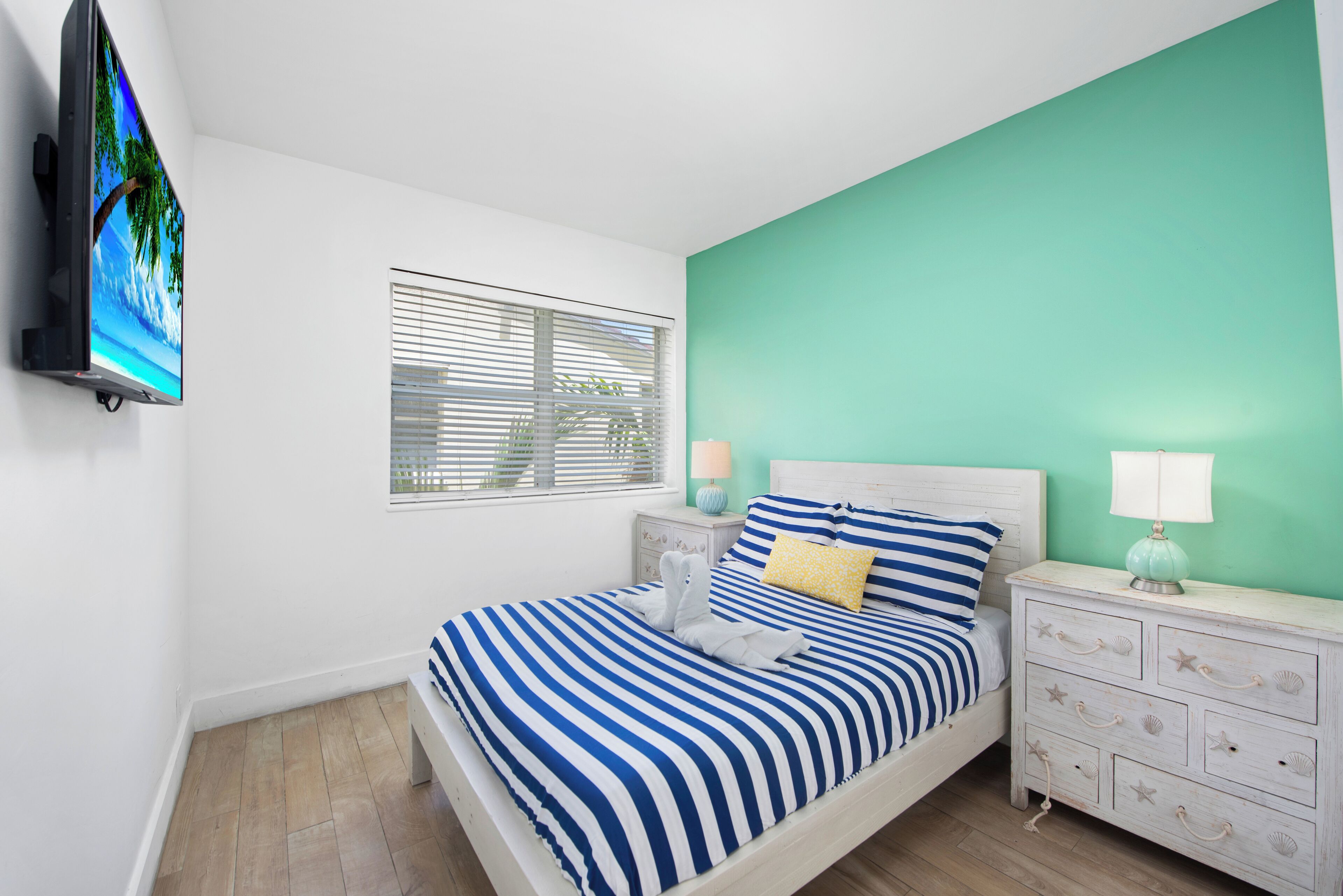 A bedroom with a bed, a nightstand, a TV mounted on the wall, and a window with blinds.
