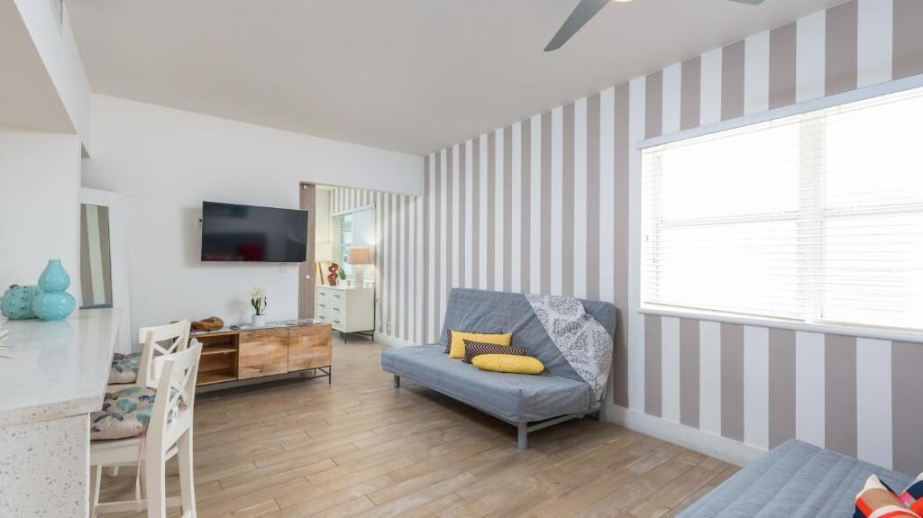 A modern living room with a sofa, a TV, a dining area with chairs, and a window with blinds.