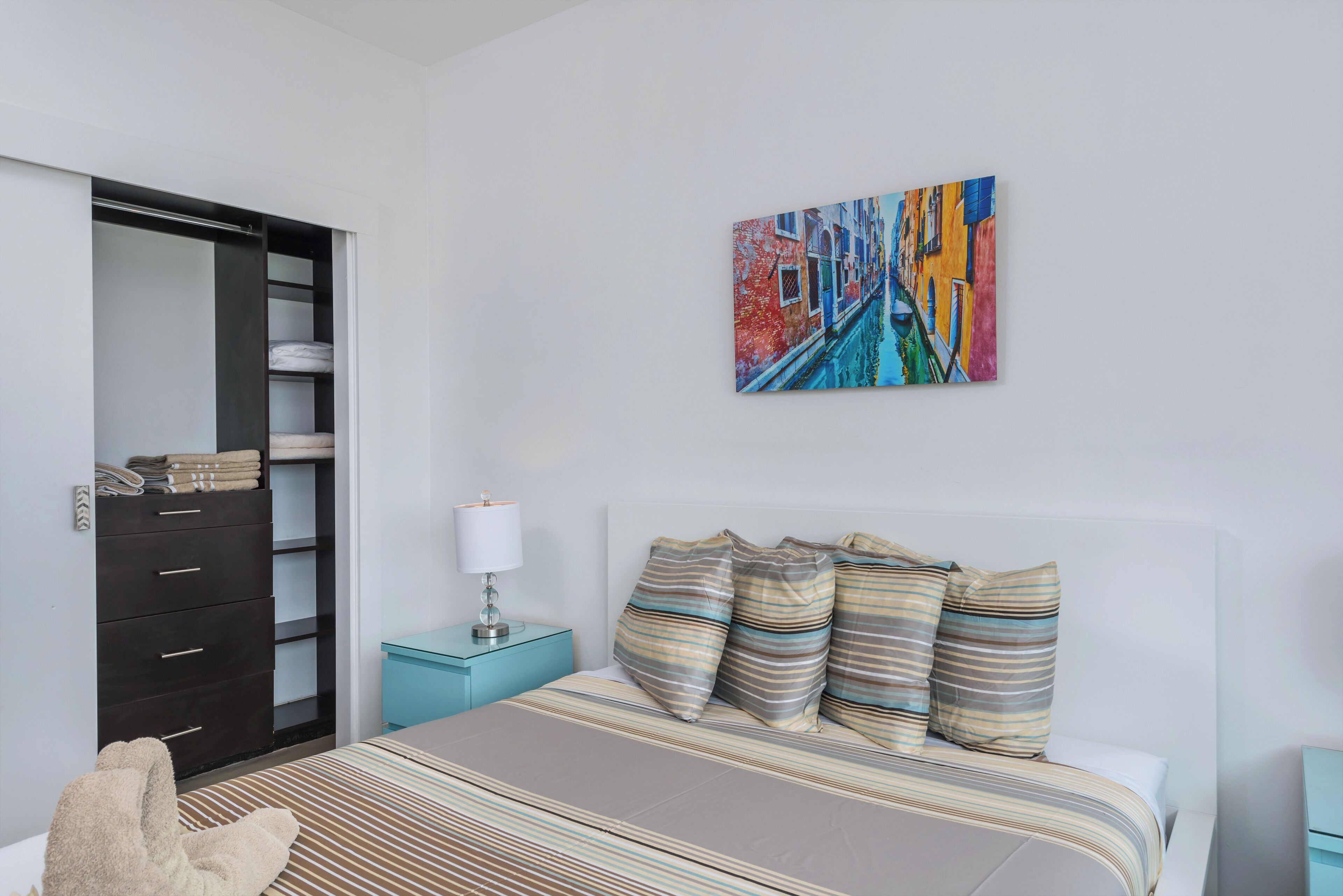 Een slaapkamer met een bed, nachtkastje, lamp, kast en een kleurrijk schilderij aan de muur.