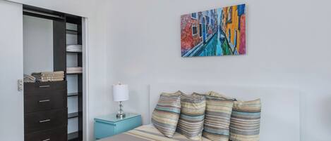 Een slaapkamer met een bed, nachtkastje, lamp, kast en een kleurrijk schilderij aan de muur.