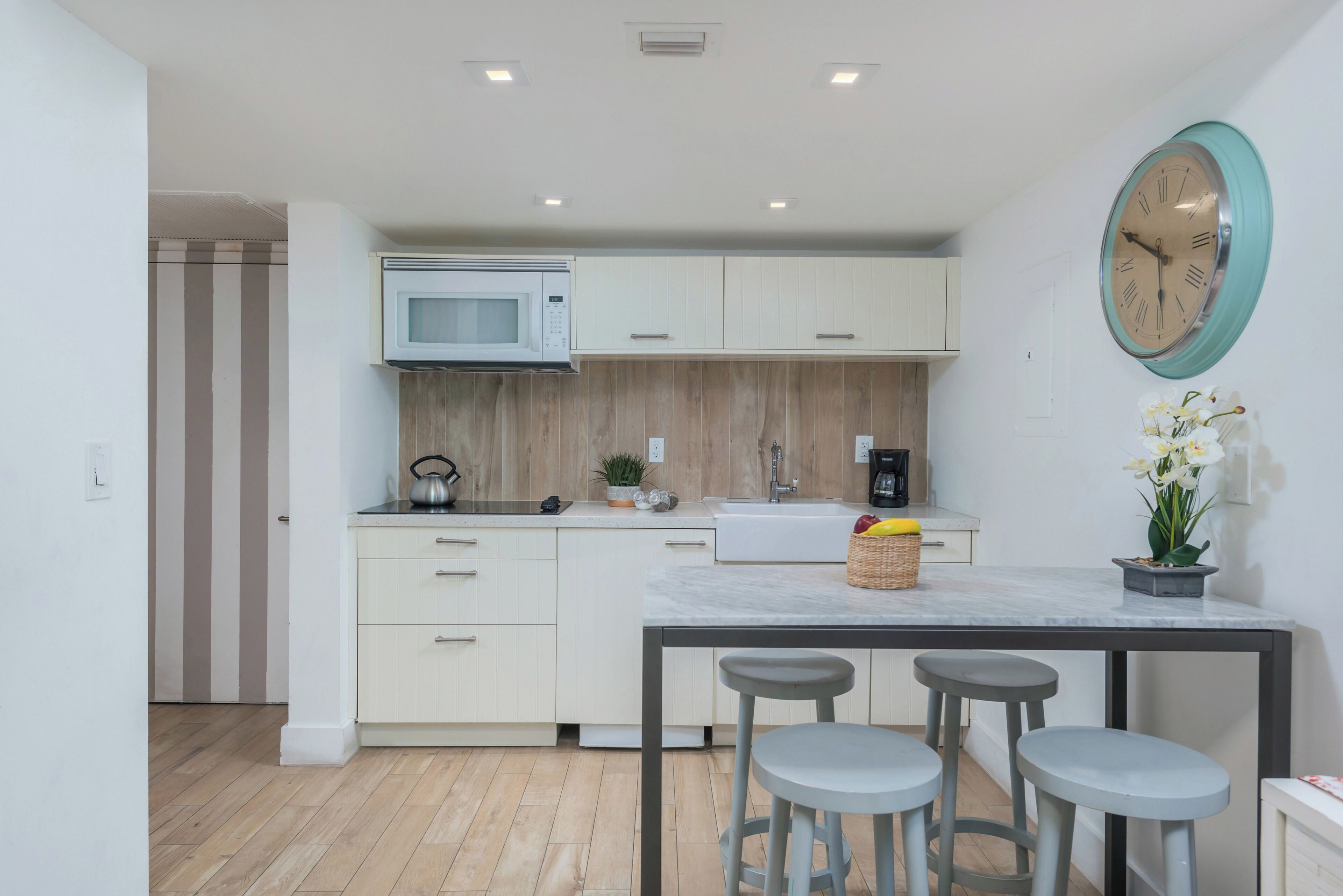 Cocina moderna con armarios blancos, microondas, fregadero y un reloj en la pared. Hay tres taburetes en una pequeña mesa.