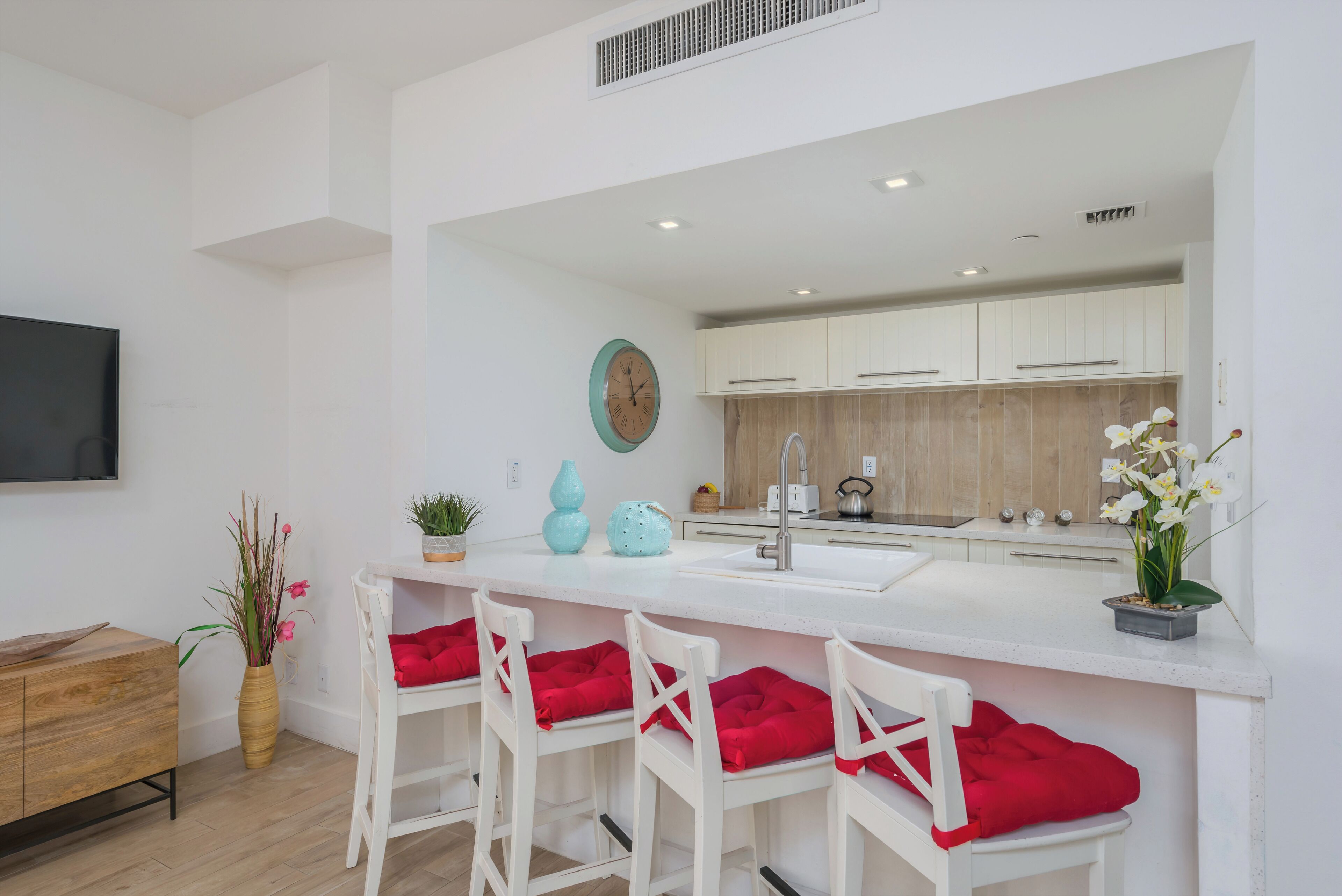 A modern kitchen with white cabinets, a large island with bar stools, a flat-screen TV, and decorative plants.