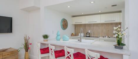A modern kitchen with white cabinets, a large island with bar stools, a flat-screen TV, and decorative plants.
