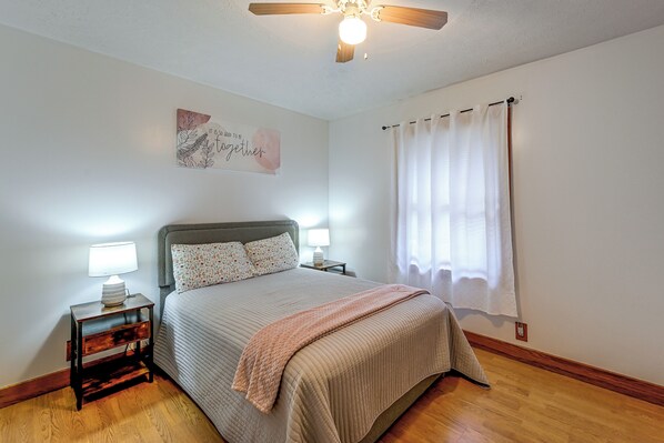 A bedroom with a bed, nightstands, a ceiling fan, a window with curtains, and a wall art piece.